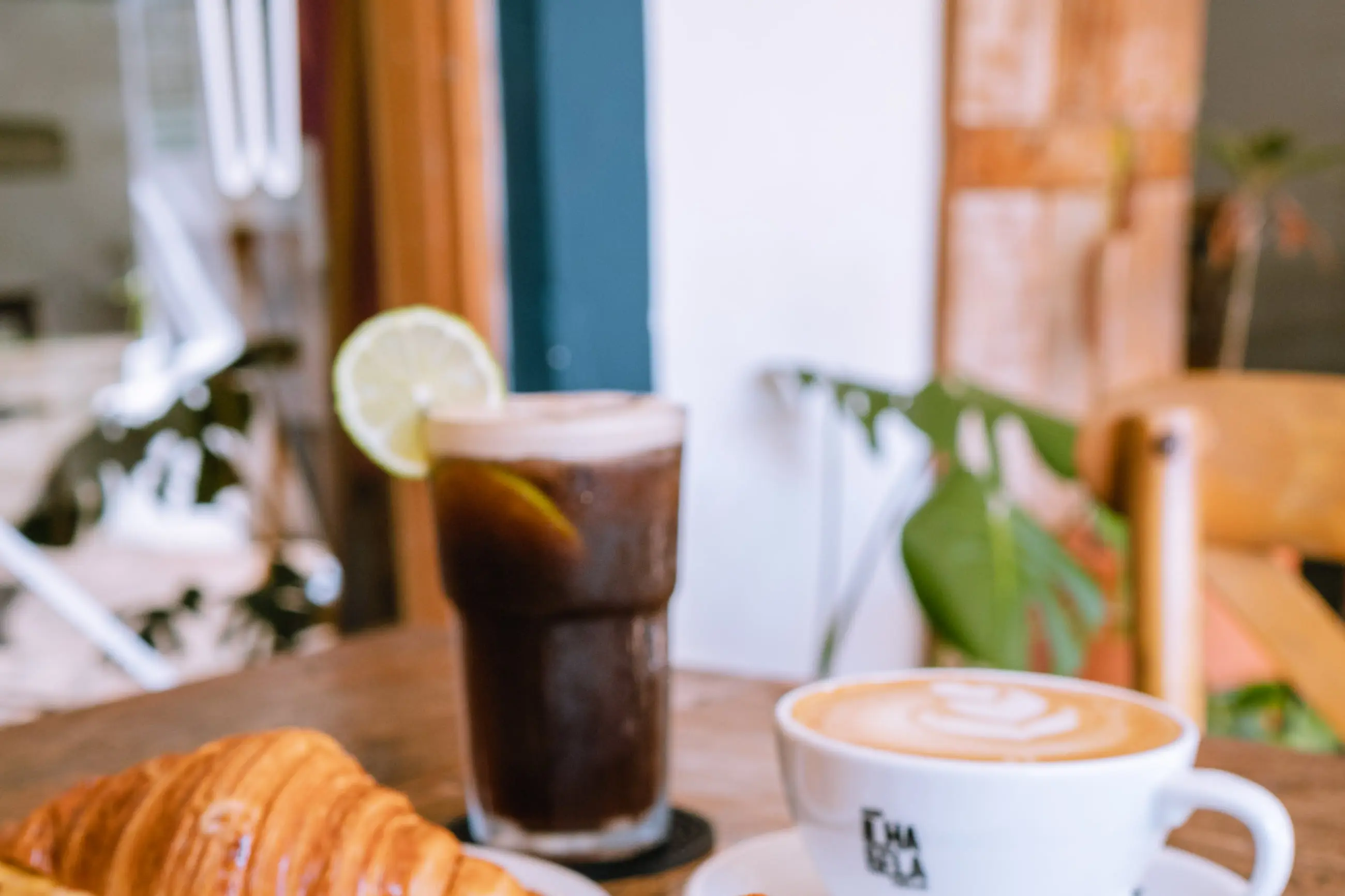Croissant, cappuccino e café gelado servidos lado a lado na mesa de madeira da casa.