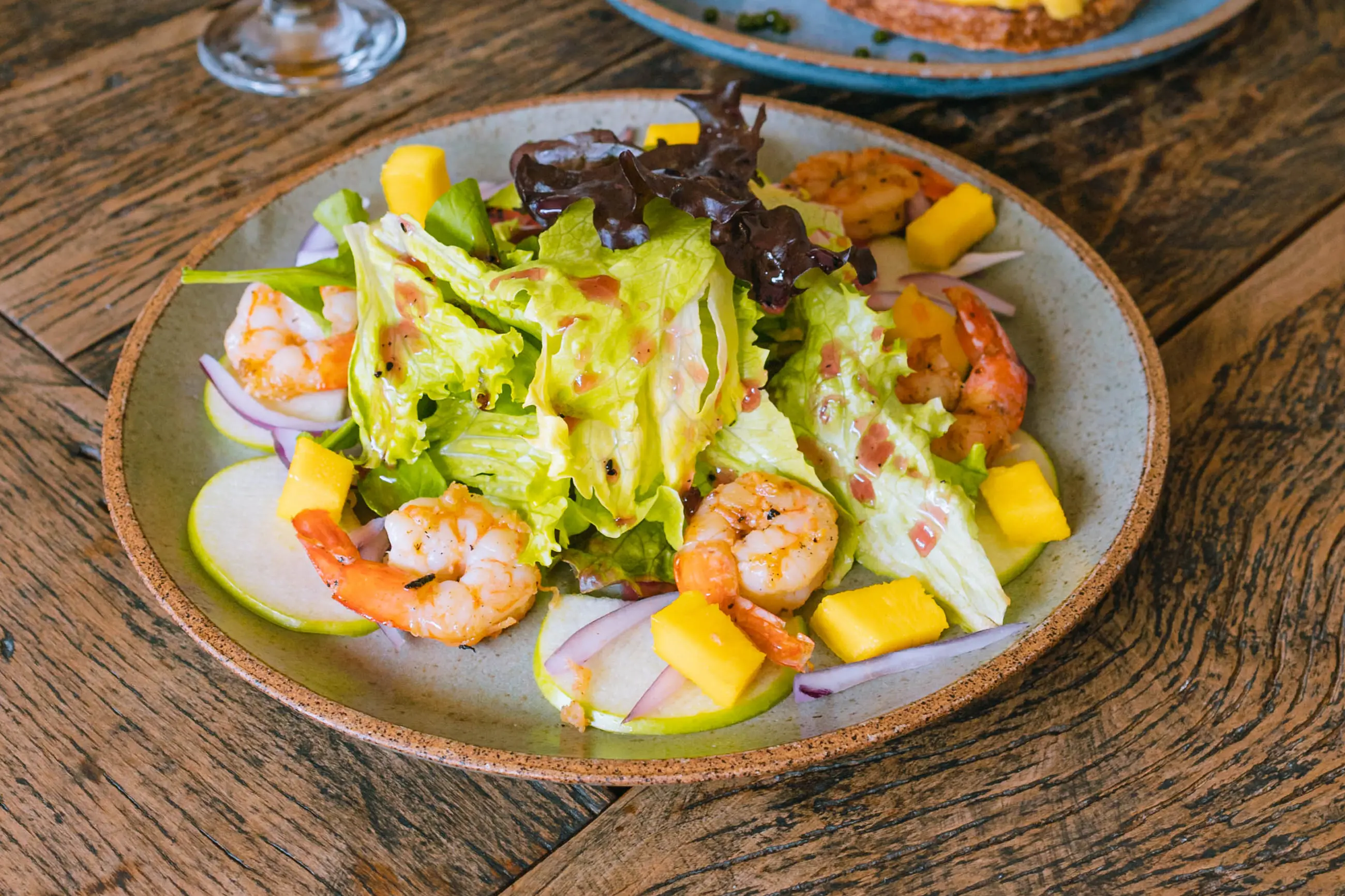 Salada com camarão, manga e folhas em prato de cerâmica, servida na mesa de madeira.