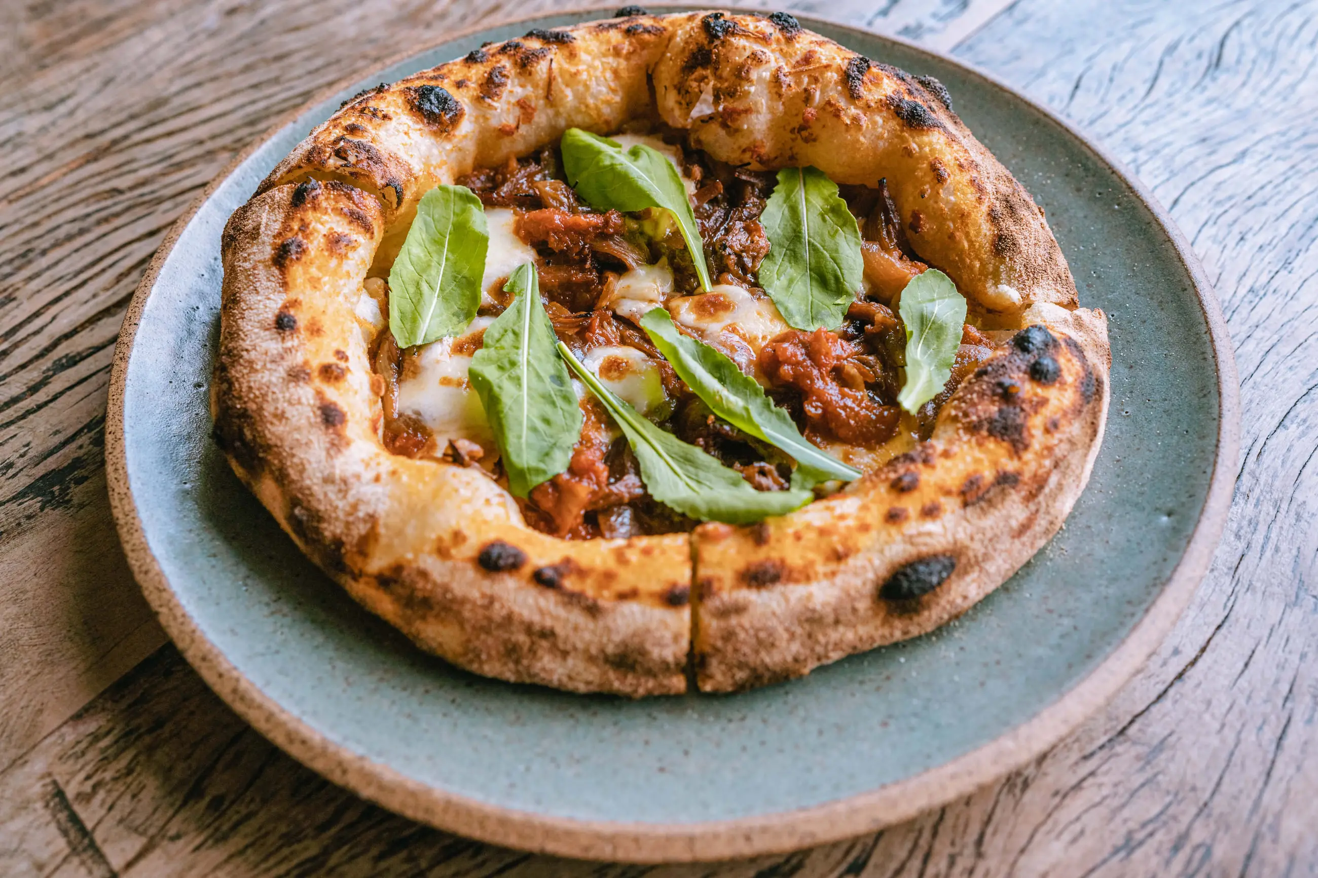 Pizza inteira com folhas frescas e borda alta de forno, servida em prato de cerâmica.