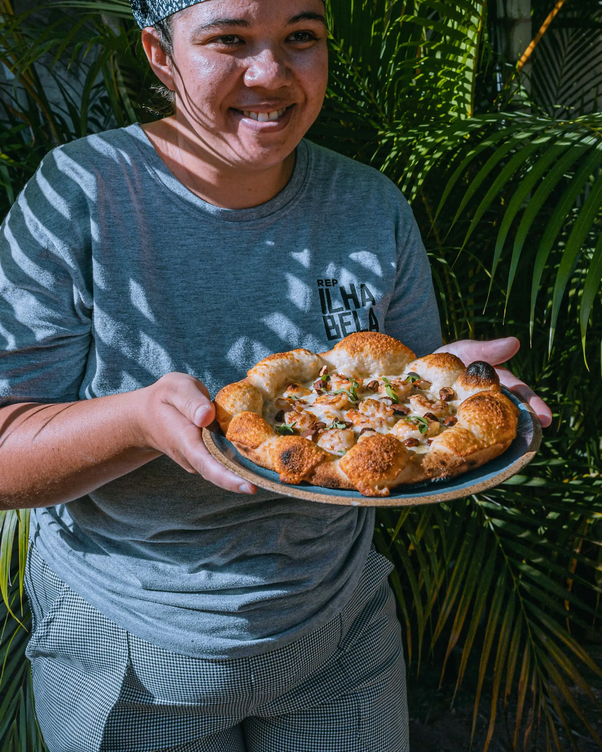Cozinheira da casa segurando, sorrindo, uma pizza que acabou de sair do forno.