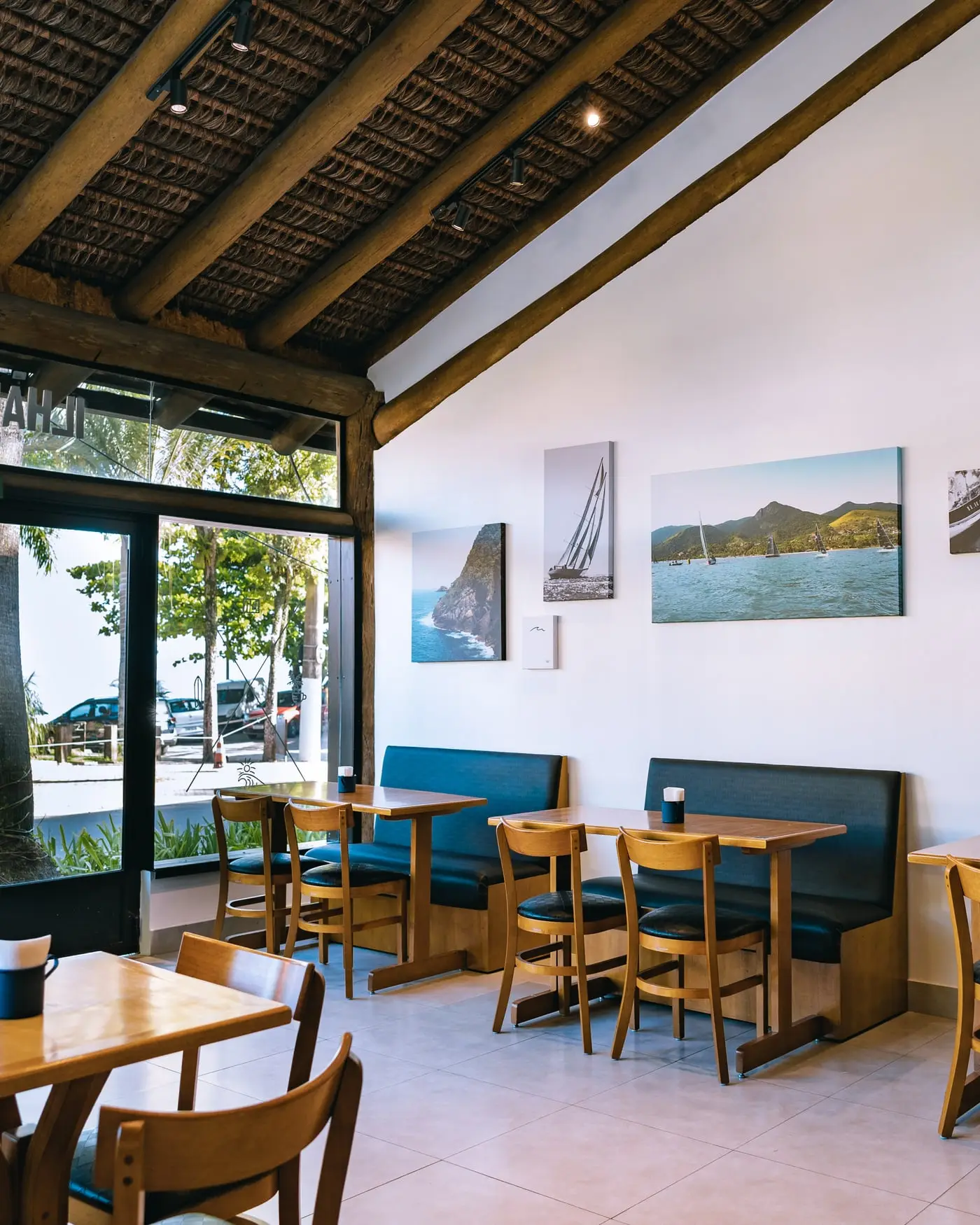 Salão da DownTown vazio de manhã: teto de galhos sobre vigas de madeira, fotografias da ilha na parede branca e mesas de madeira com bancos azuis.