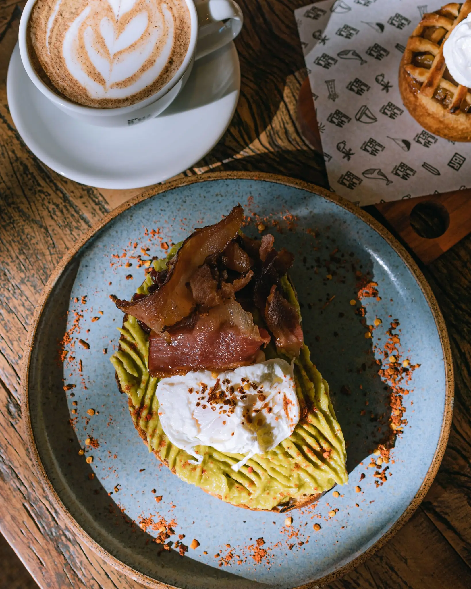 Torrada de avocado com bacon e ovo poché, polvilhada com pimenta, num prato azul; ao lado, um cappuccino na xícara da casa e uma torta de maçã sobre o papel estampado da marca.