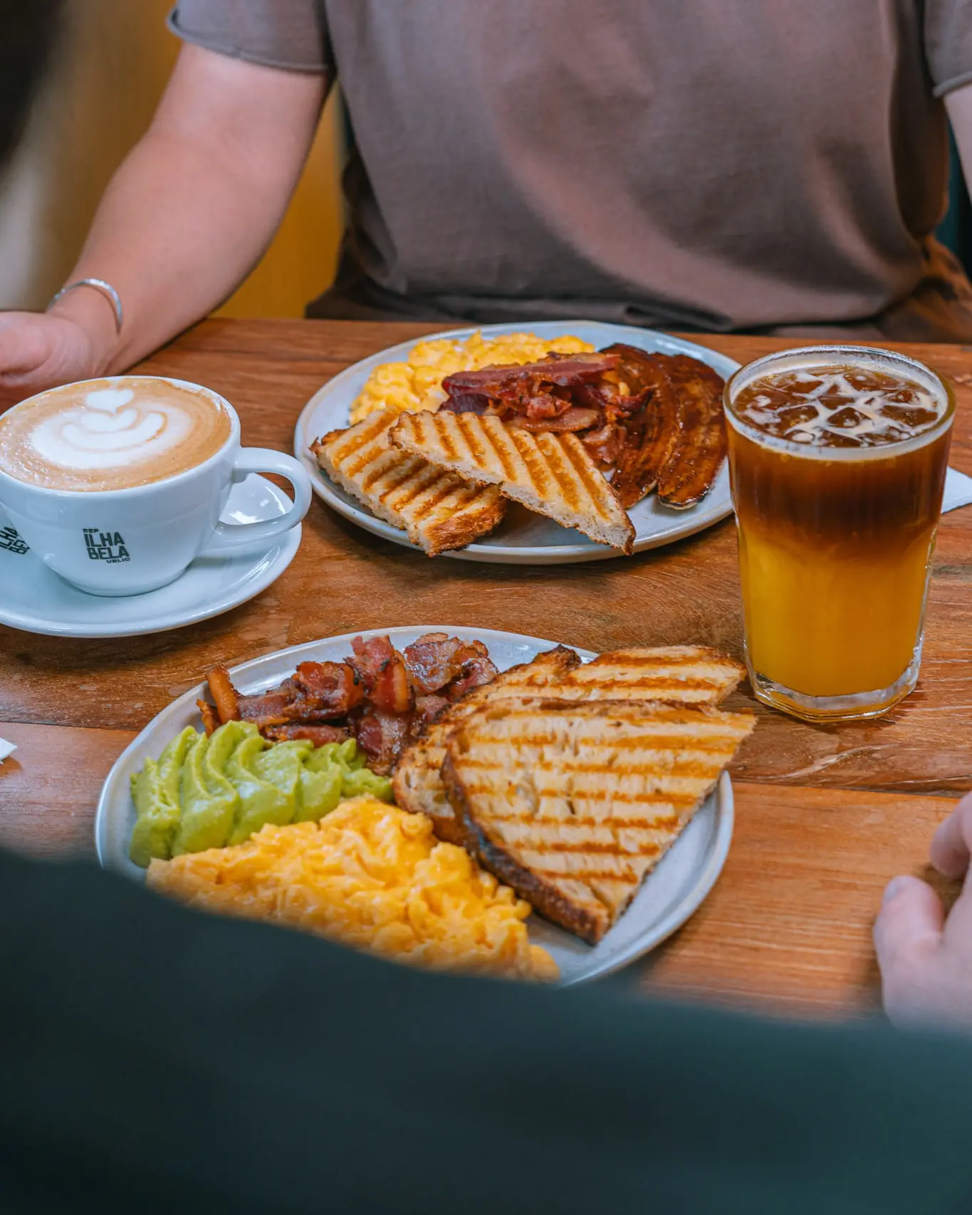 Mesa de brunch para dois: cappuccino na xícara da casa, dois pratos com ovos mexidos, bacon, avocado e pão na chapa, e um copo de café com suco de laranja.