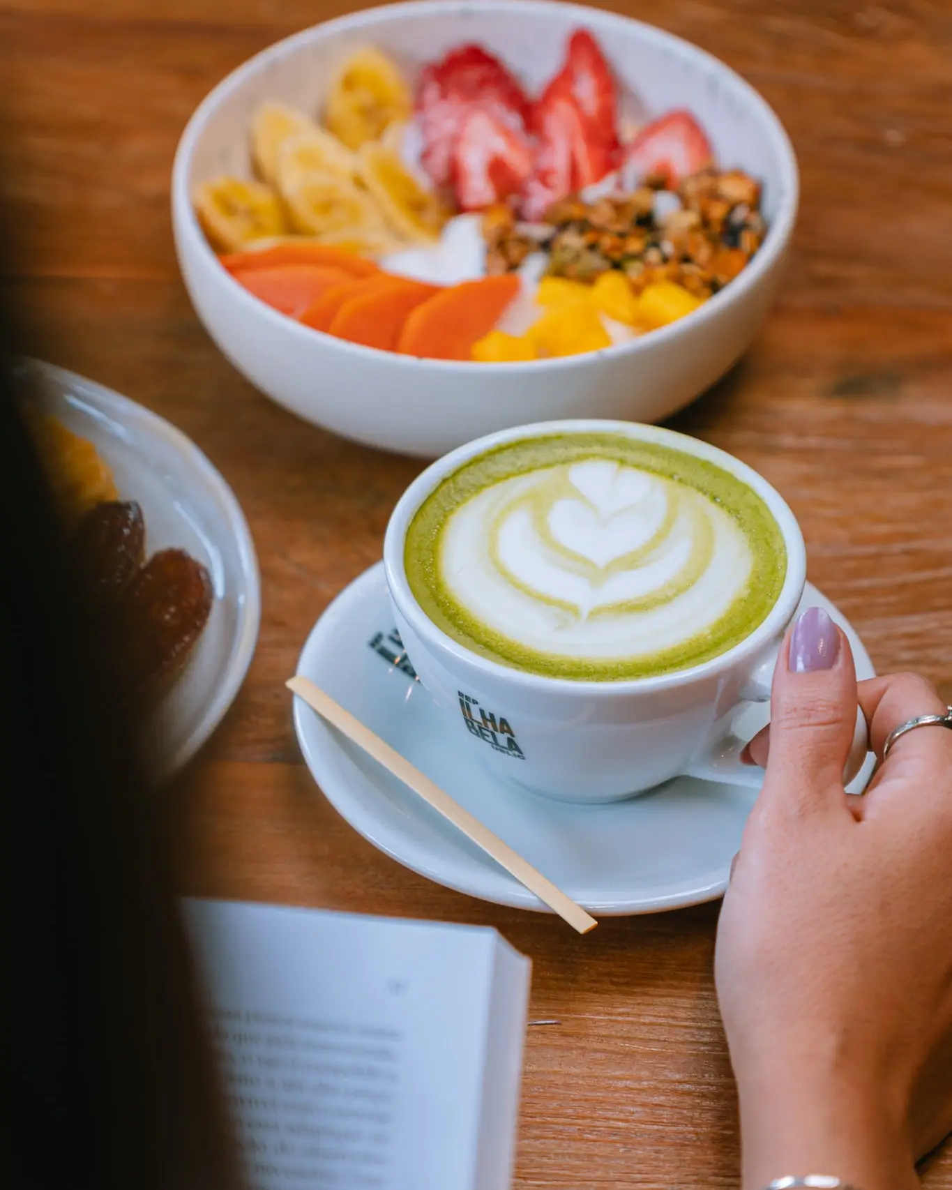Matcha latte na xícara da casa, com a arte desenhada no leite, ao lado de uma tigela de frutas com granola e de um livro aberto sobre a mesa de madeira.