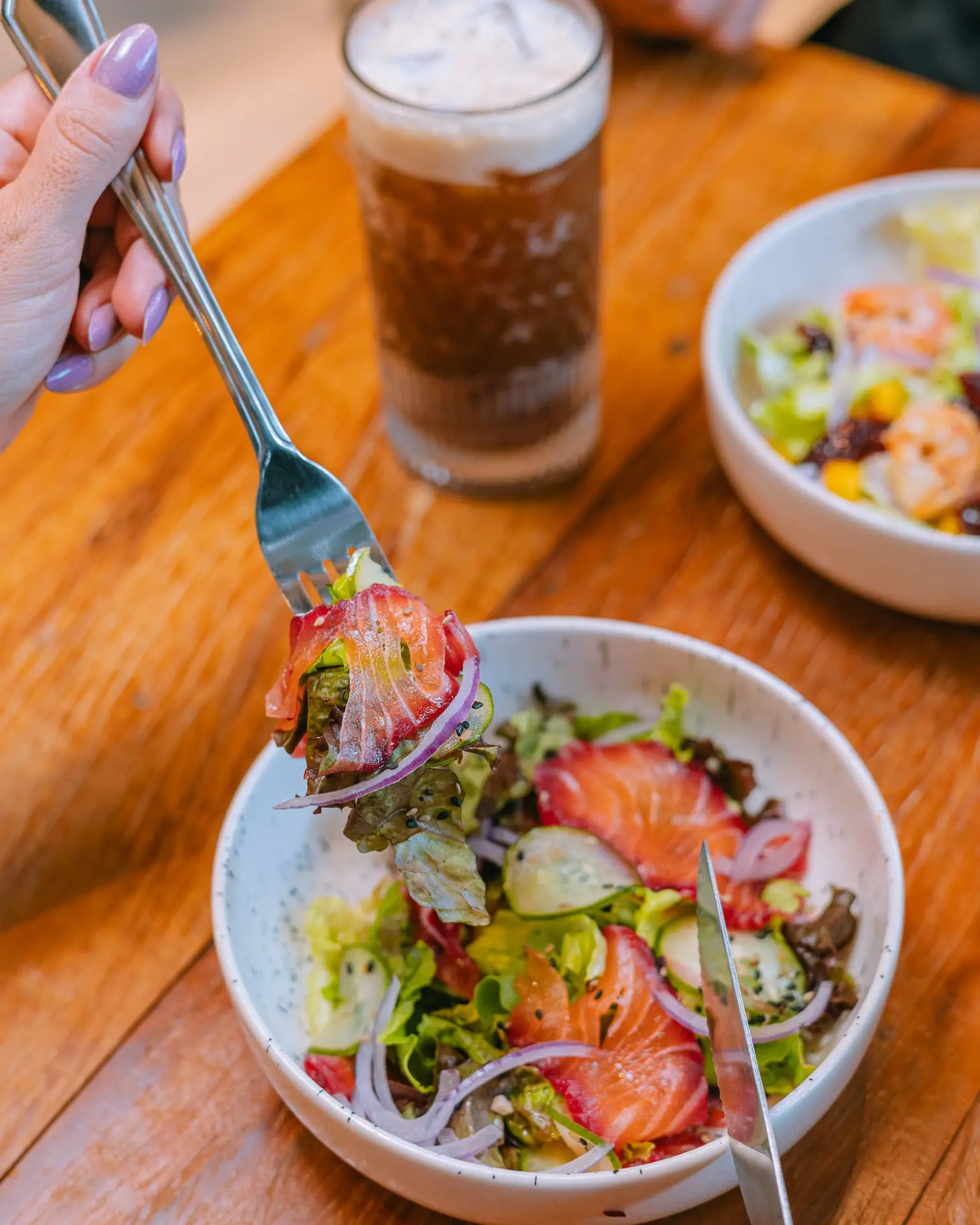 Salada de folhas com salmão, morango, pepino e cebola roxa, com o garfo levantando uma porção e uma segunda tigela ao fundo.