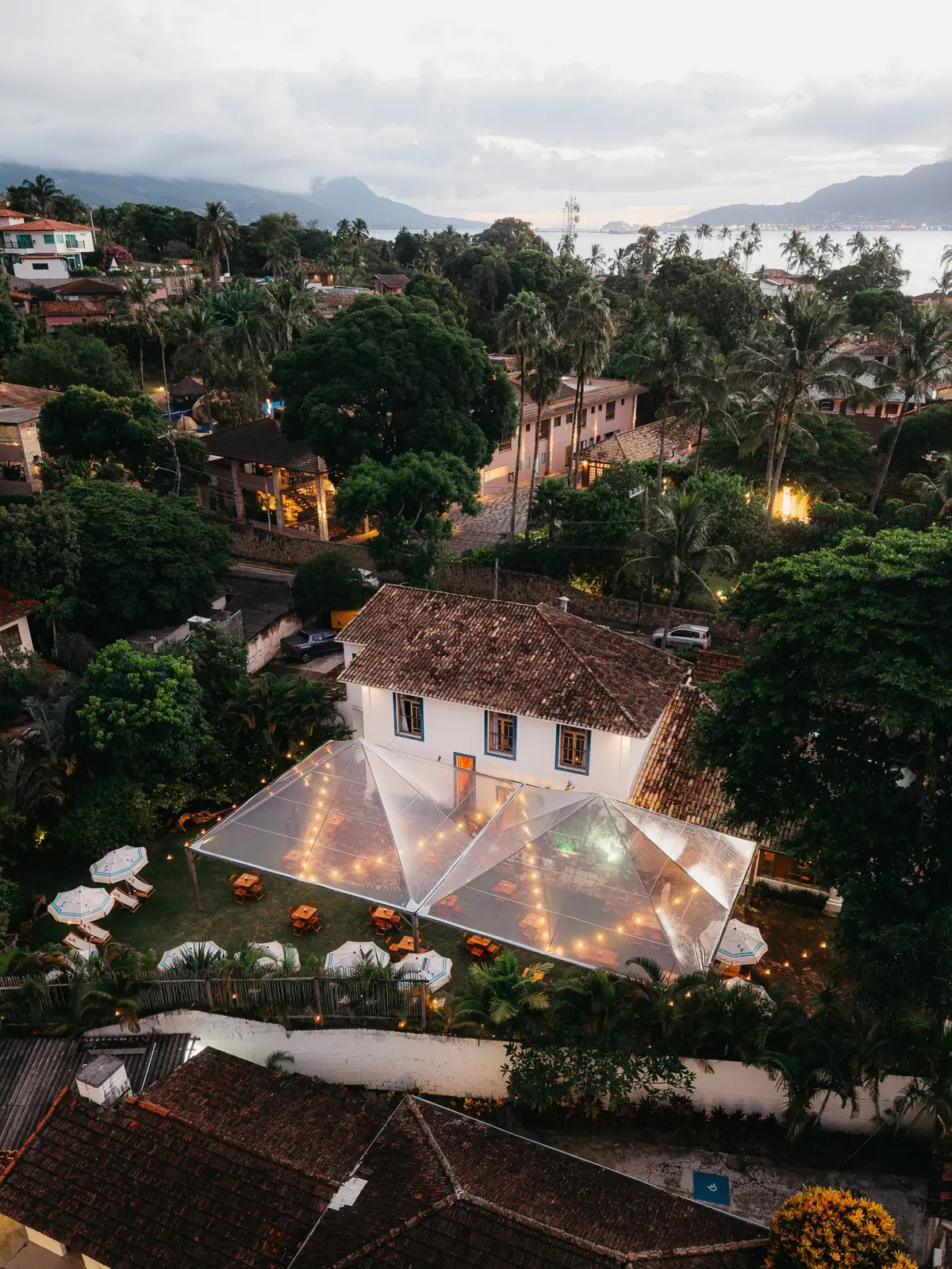 Vista aérea do casarão colonial da Garden no fim da tarde, com o toldo transparente montado sobre o jardim e o canal de São Sebastião ao fundo.