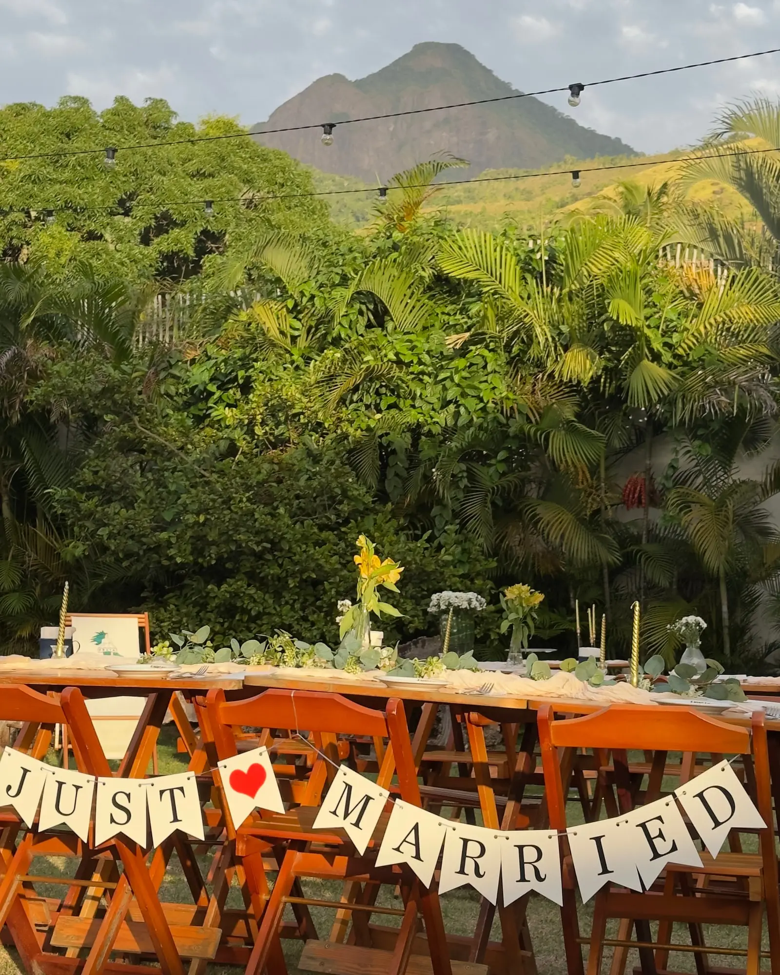 Mesa comprida posta para um casamento no jardim da Garden, com a faixa JUST MARRIED nas cadeiras e o Pico do Baepi ao fundo.