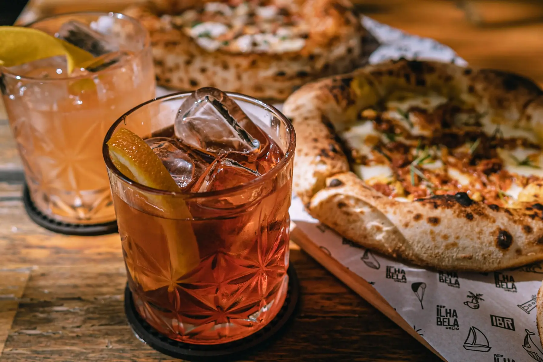 Dois drinks com gelo e casca de laranja ao lado de duas pizzas recém-saídas do forno, sobre a mesa de madeira e o papel estampado da marca.