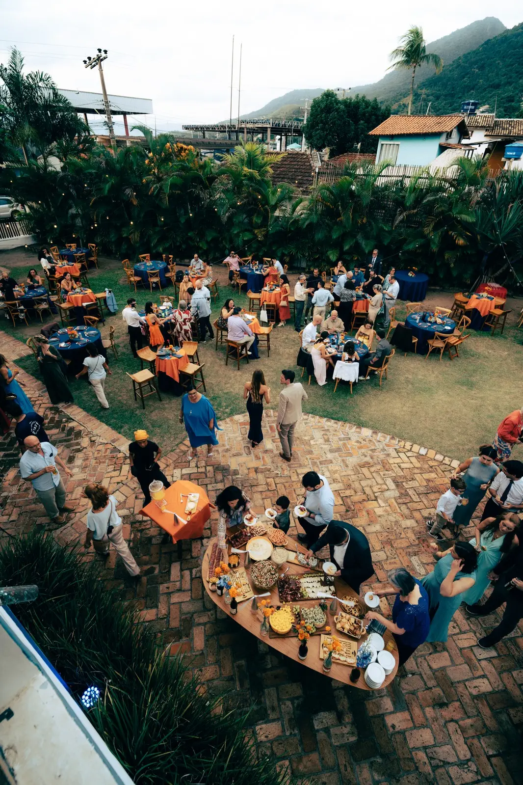 Vista de cima do jardim durante um casamento: mesas redondas postas no gramado e uma ilha de comida sobre o piso de tijolo.