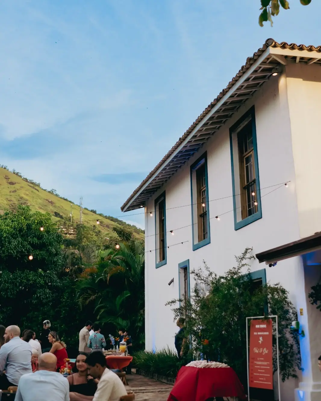 Casarão colonial de janelas azuis ao entardecer, com o telhado de telha, luzes penduradas e convidados às mesas no jardim, com o morro atrás.