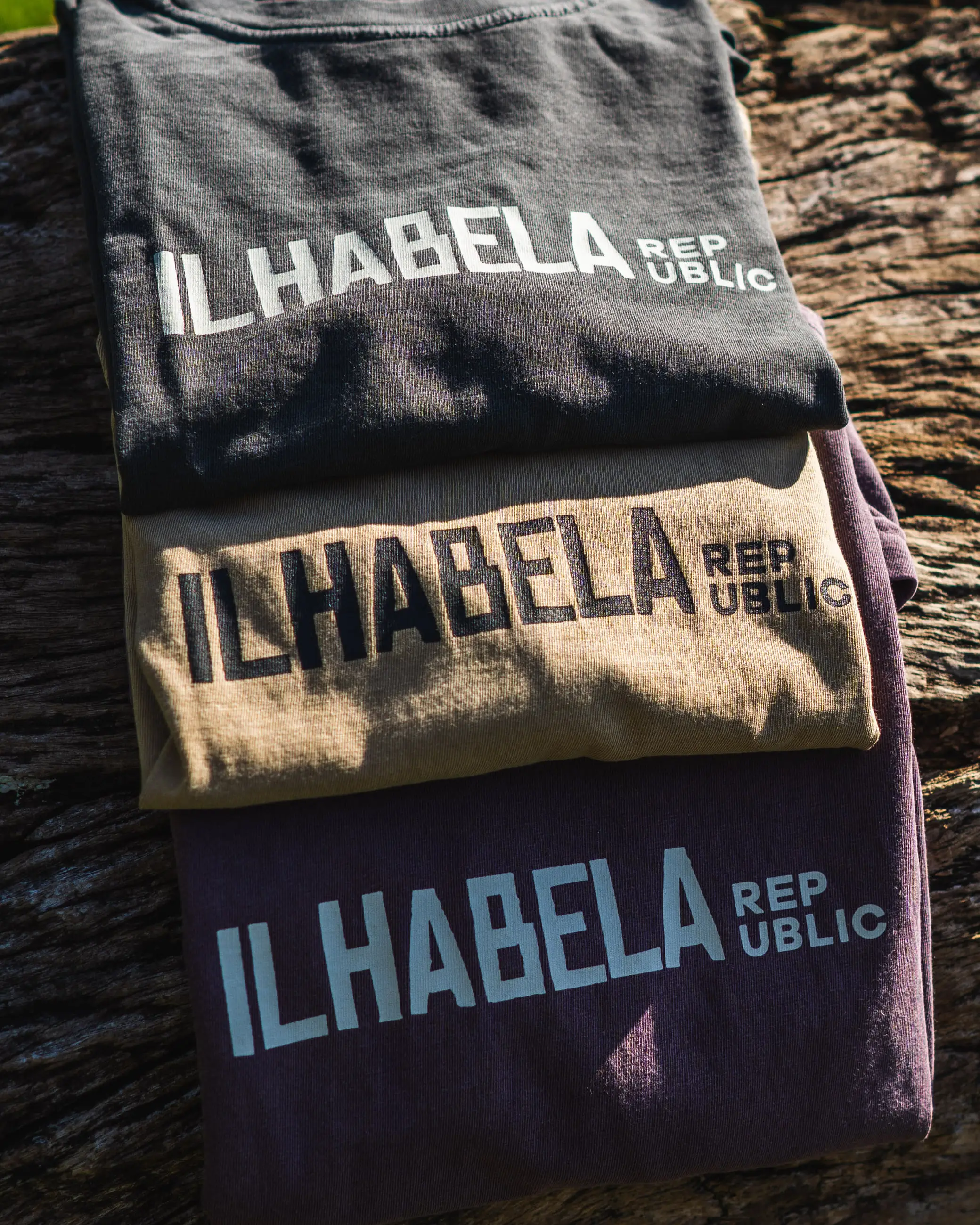 Três camisetas da Ilhabela Republic dobradas sobre um tronco — chumbo, areia e vinho, com o logotipo estampado no peito.