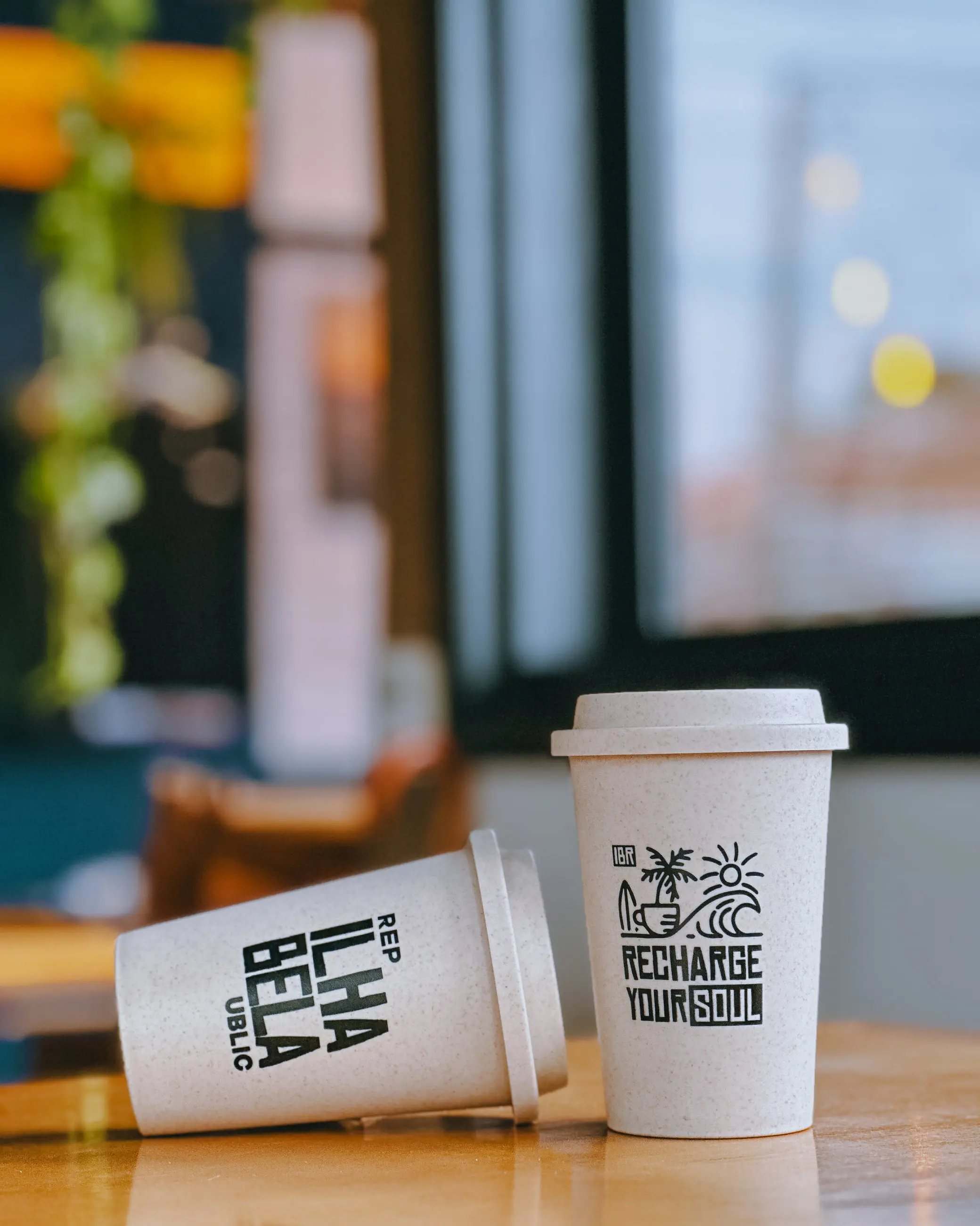 Dois copos de viagem da Ilhabela Republic sobre a mesa de madeira, um deles com o logotipo da marca.