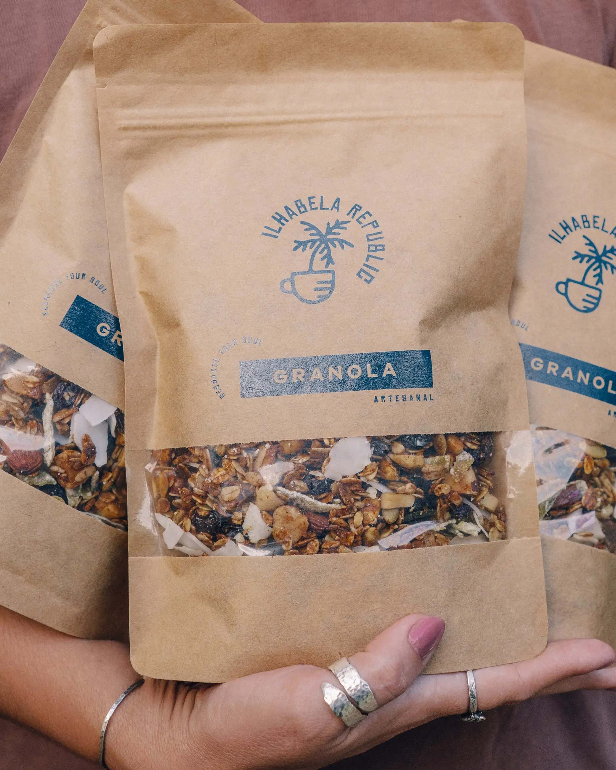 Três pacotes kraft de granola artesanal da Ilhabela Republic com janela transparente, carregados no braço.