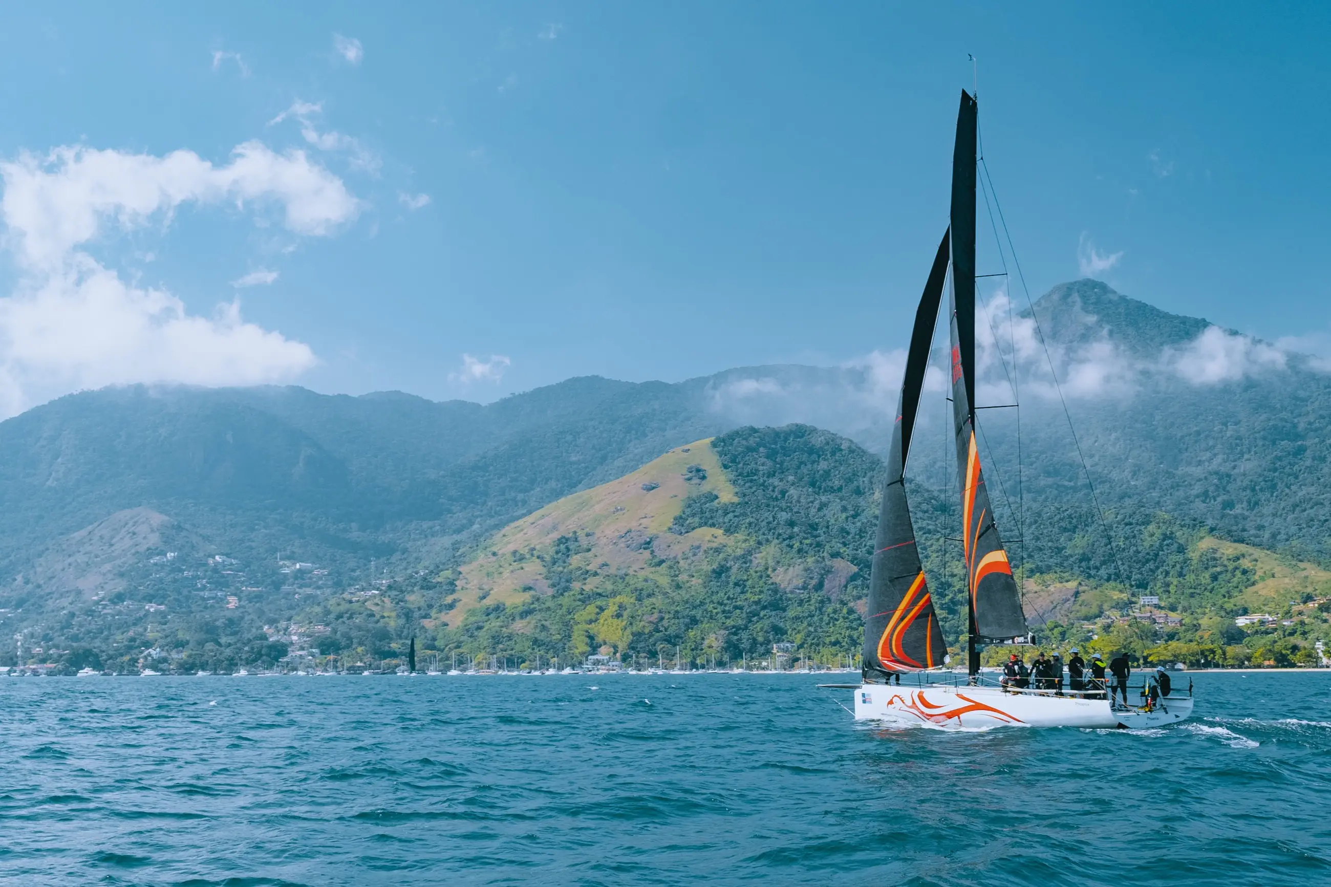 Veleiro de regata navegando no canal de São Sebastião, com a vila de Ilhabela na orla, veleiros fundeados na marina e a serra coberta de mata subindo atrás.