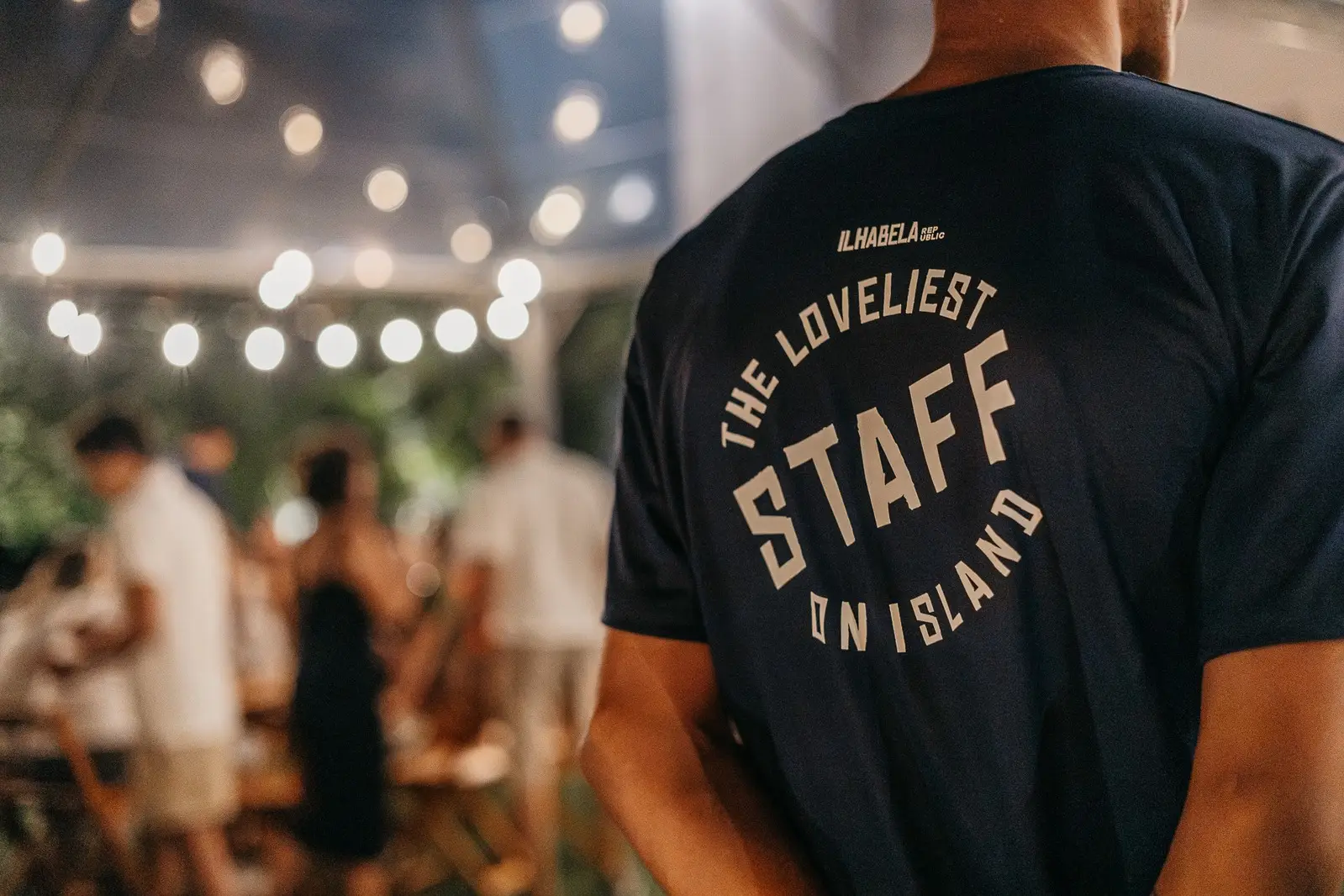 Integrante da equipe de costas, com a camiseta da casa estampada The Loveliest Staff on Island, num evento à noite sob luzes penduradas.