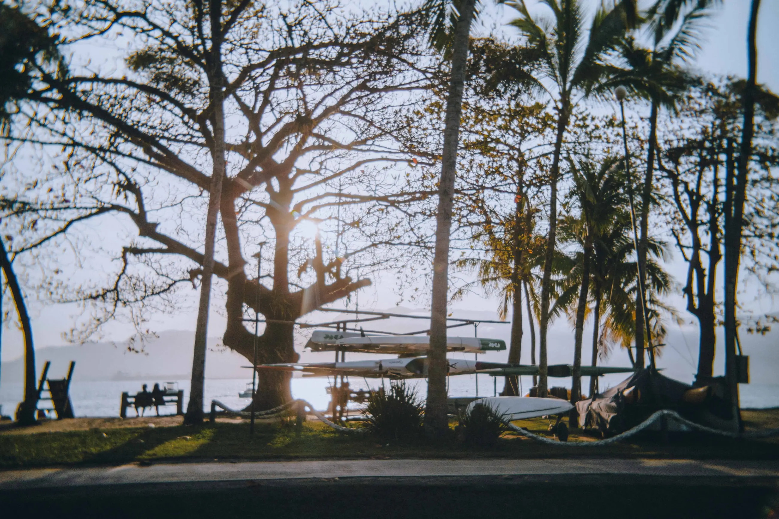 Árvores e coqueiros de uma praça na orla ao entardecer, com bancos, canoas na areia e o mar ao fundo.