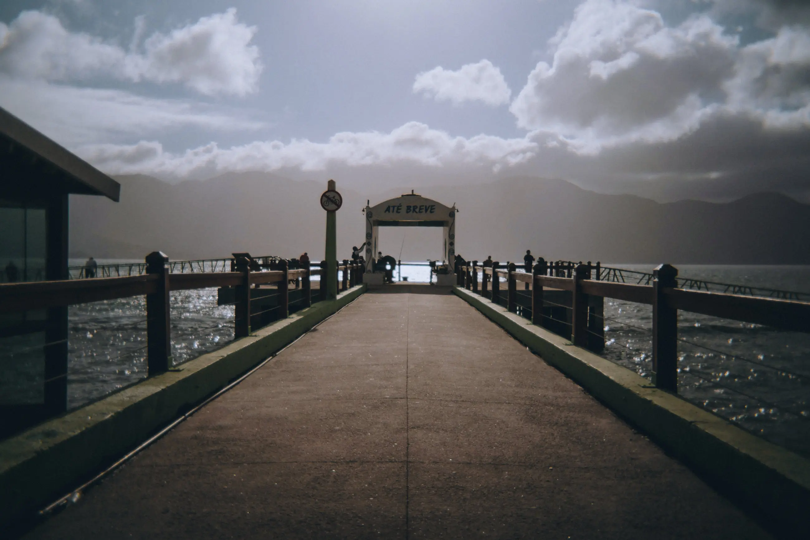 O píer de Ilhabela em dia nublado, com o deque de madeira seguindo reto até o portal sobre a água.