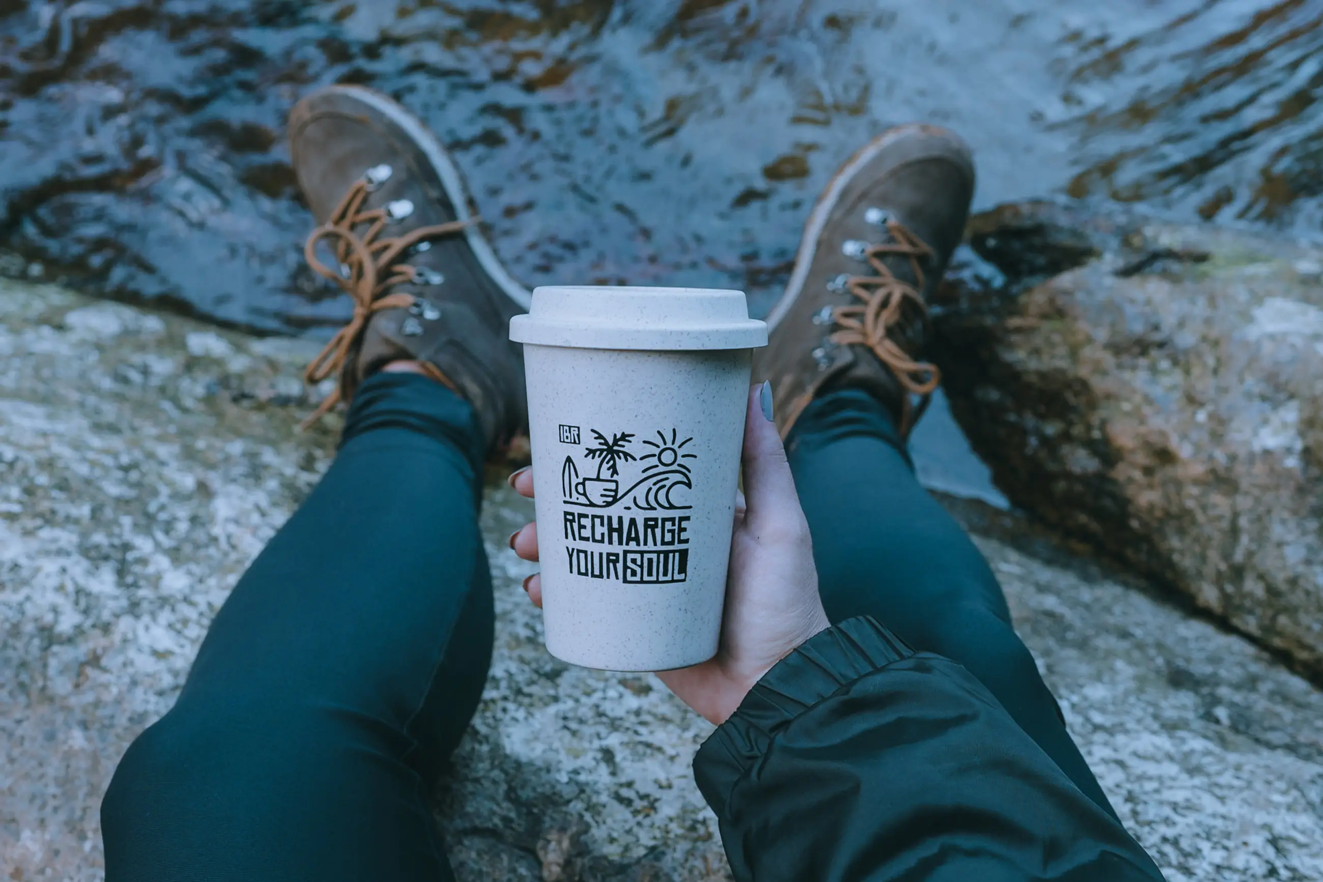 Copo de viagem da Ilhabela Republic apoiado na pedra de um rio, no meio da trilha, com as botas de quem parou para o café.