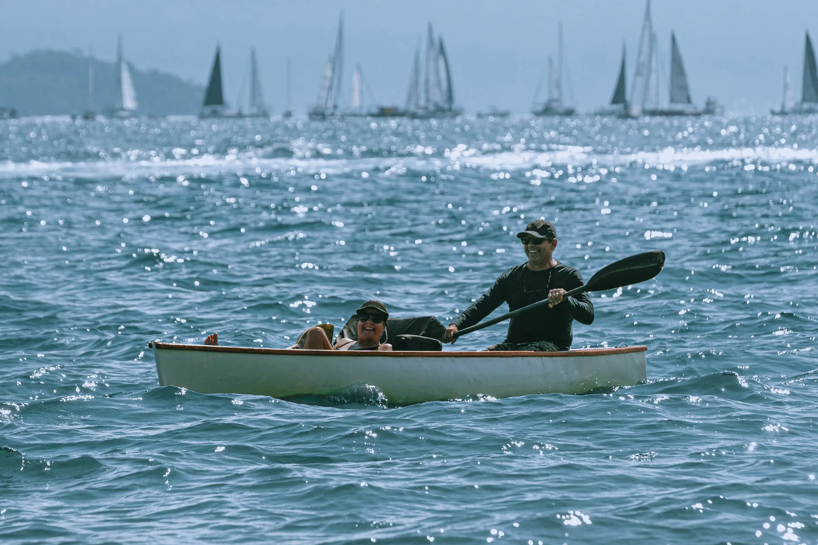 Homem remando uma canoa no canal, com a frota de veleiros da regata alinhada no horizonte atrás.